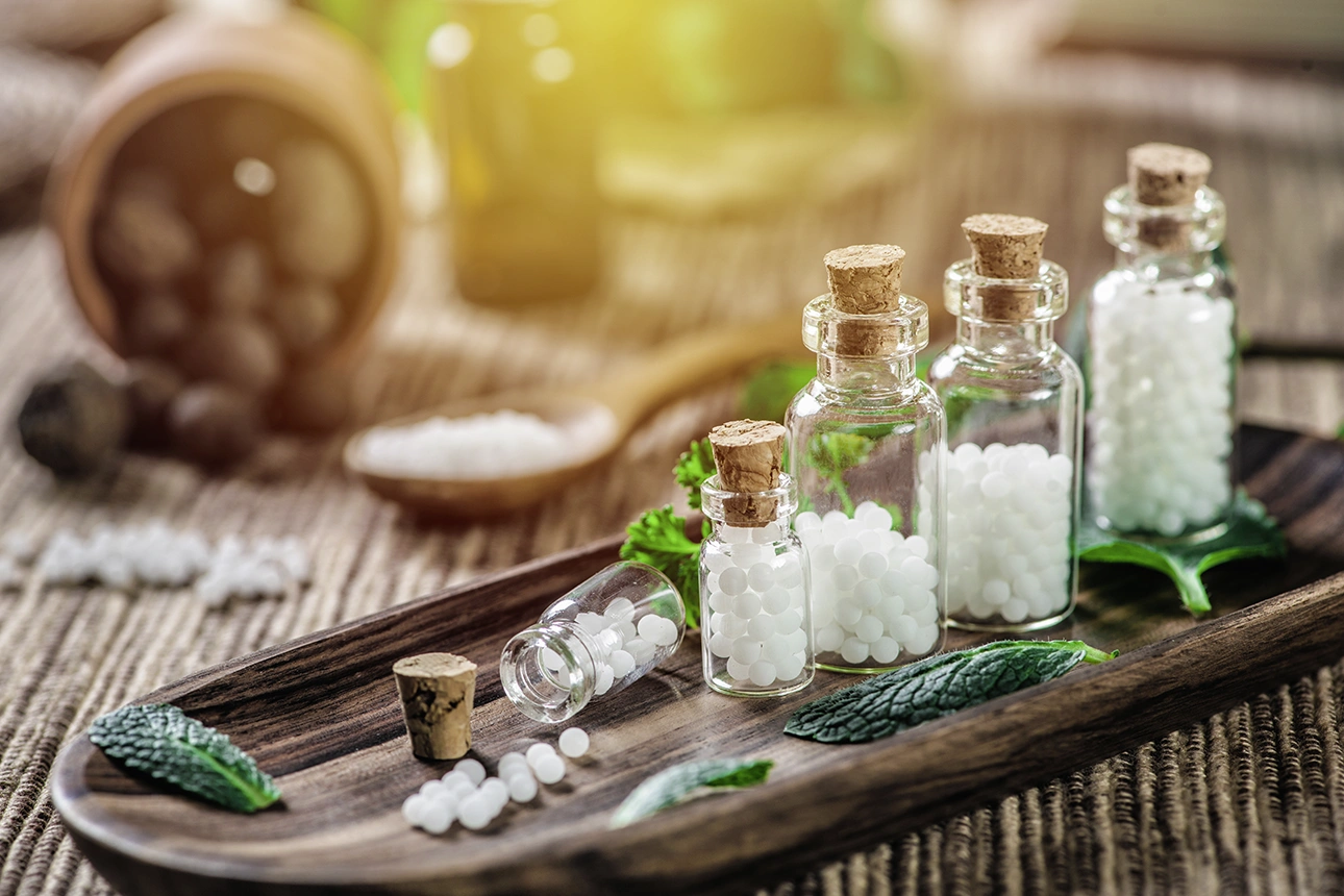 Auf einem Holzbrett stehen mehrere kleine Glasfläschchen mit Korkverschlüssen, die mit weißen, Kügelchen gefüllt sind. Diese Kügelchen sind typisch für homöopathische Globuli.