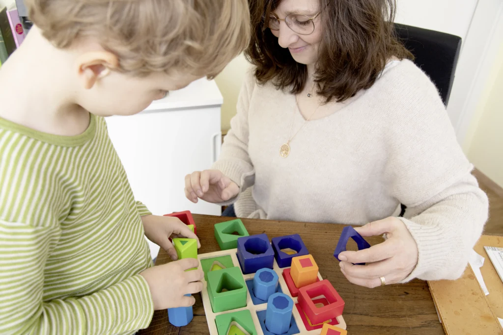 ChatGPT: Kerstin und ein kleiner Patient führen eine ergotherapeutische Übung mit Holzformen durch, bei der motorische und kognitive Fähigkeiten spielerisch gefördert werden.
