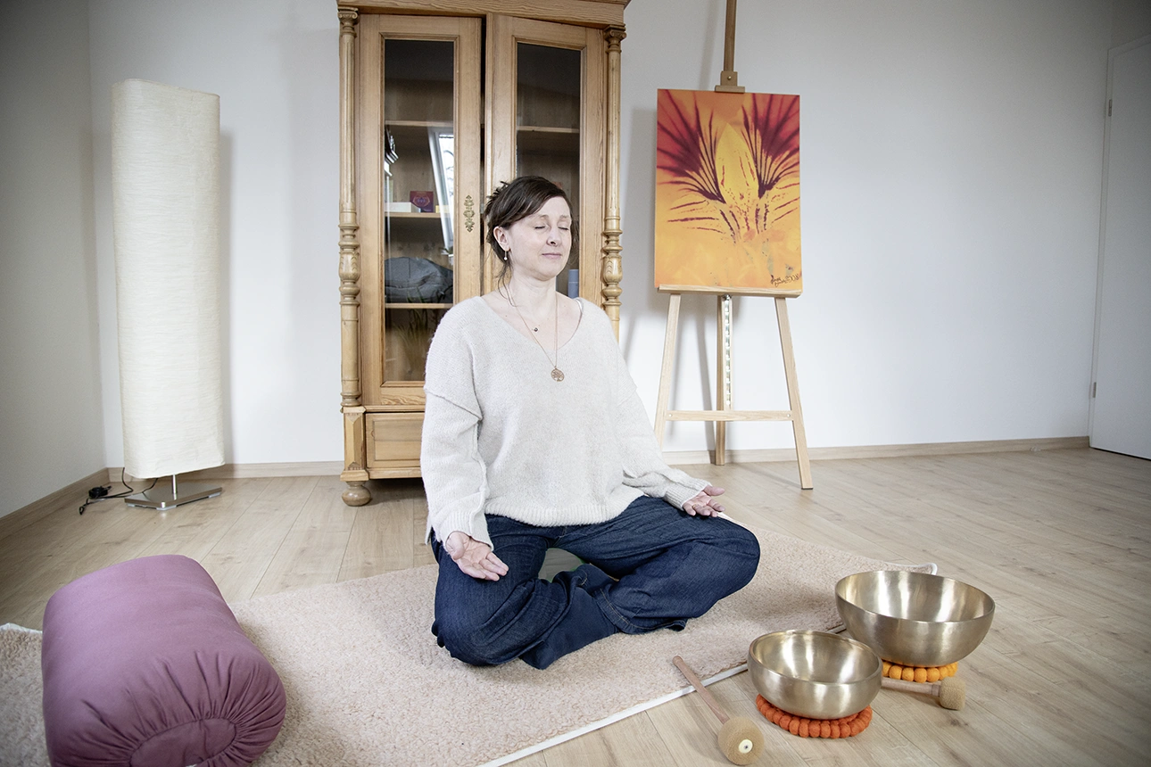 Kerstin Winner sitzt auf einem Meditationskissen und meditiert auf einer Matte. Vor ihr stehen Klangschalen.